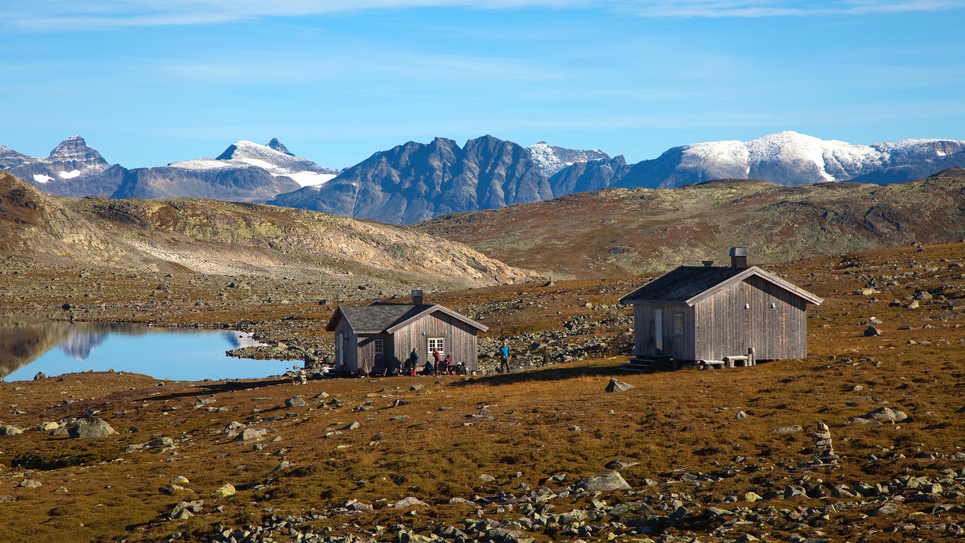 To hytter i åpent fjell ved et vann, badet i høstsol. Høye fjelltopper med nysnø på i bakgrunnen.