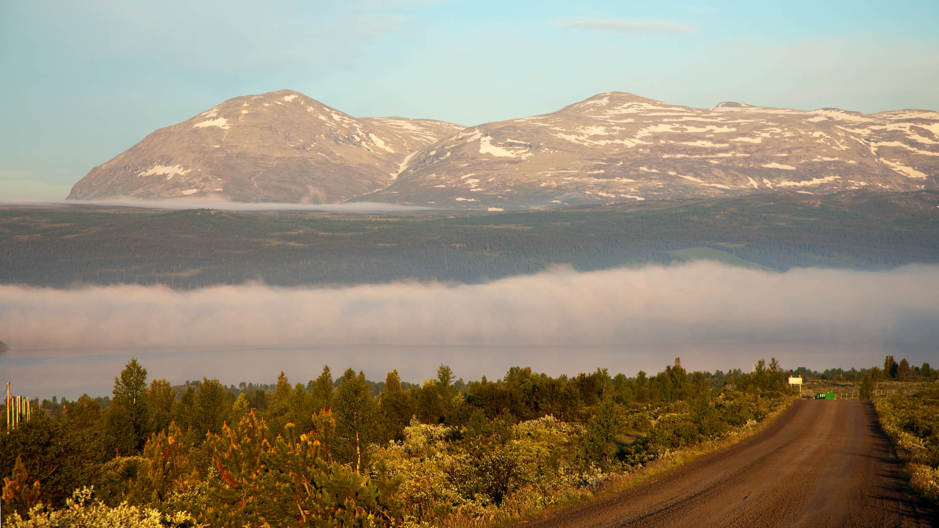 På en stølsslette med buskvegetasjon i tidlig morgensol, mens det enda ligger morgentåke i en forsenkning. En fjellkjede reiser seg mektig i bakgrunnen.