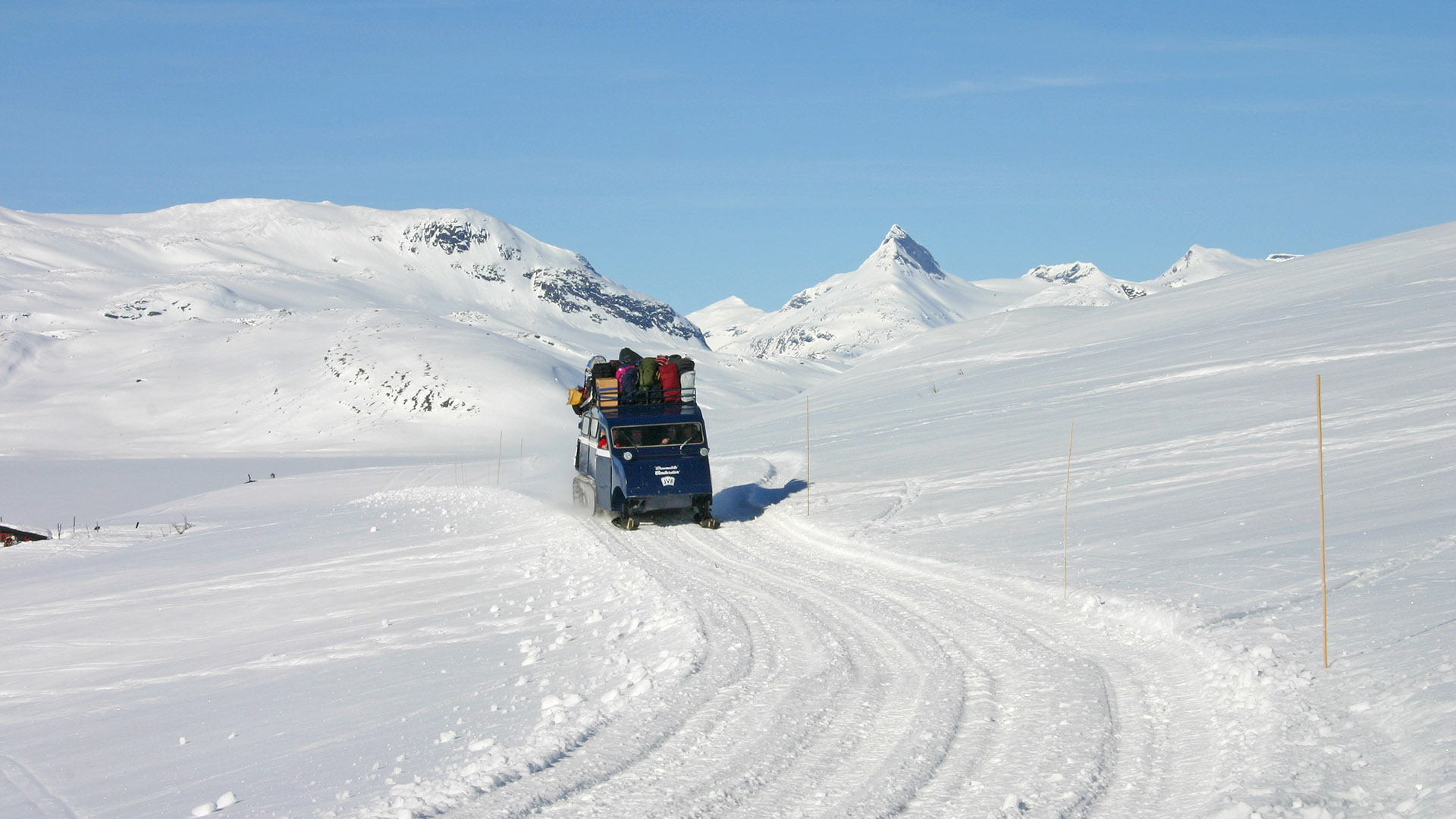 Beltebil sett forfra på oppkjørte løyper en skyfri vinterdag med Jotunheimen i bakgrunnen.